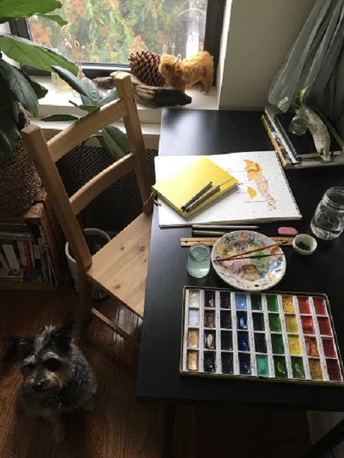An image of author and illustrator Tessa Allen's wokspace, showing paints and a work in progress on the table and her dog.