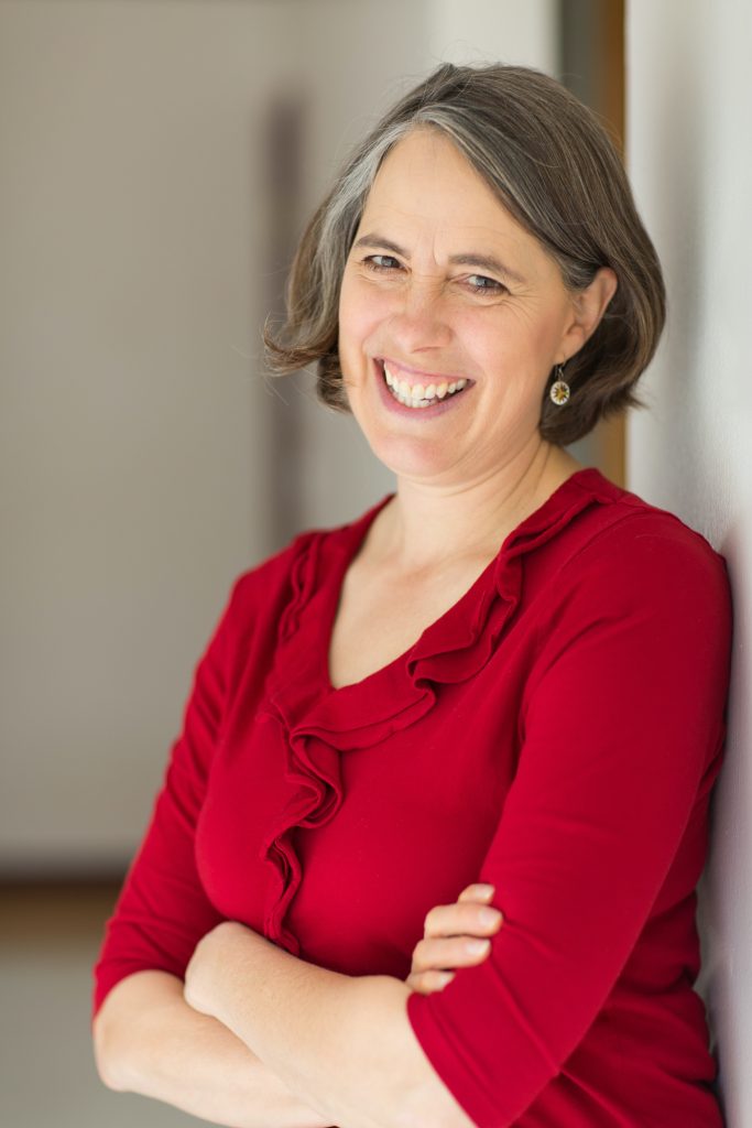 Image Description: Author Annette Bay Pimentel smiles leaning against a way wearing a red shirt.