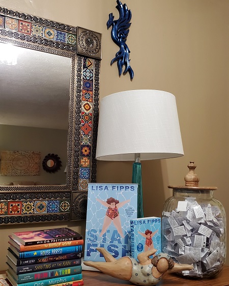 The space where author Lisa Fipps does virtual events. Pictured is a table with a mirror, a stack of books for young people, copies of Starfish and a figurine of a swimmer, and a jar filled with quotes on slips of paper.