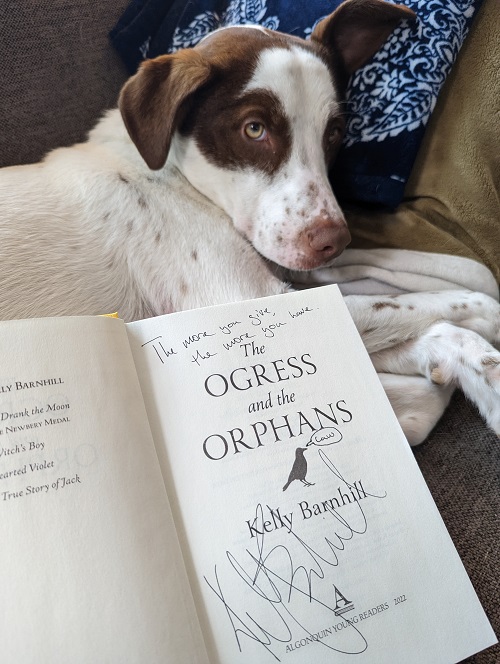 The title page of The Ogress and the Orphans, signed by the author, Kelly Barnhill, with the message, "The more you give the more you have." Kelly's brown-and-white dog rests near the open book. 