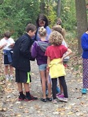 Melissa Stewart with school-age children in the forest, with leaves scattered around their feet. The children are gathered around Melissa.