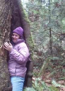 Melissa Stewart stands halfway into a large hole in a tree trunk in a forest. She is wearing a purple jacket and hat.