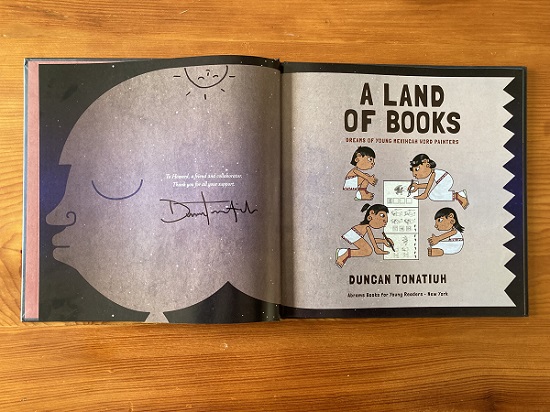 The title page spread of A Land of Books, signed by the author and illustrator Duncan Tonatiuh, with an image of an upside-down smiling sun above the sginature.