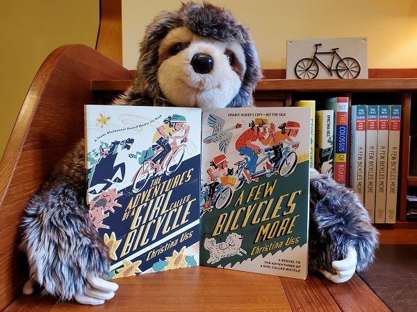 Christina Uss's stuffed sloth, holding copies of The Adventures of a Girl Called Bicycle and A Few Bicycles More.