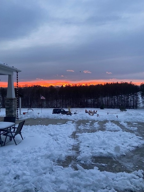 Author Amber McBride's snowy backyard at dusk with a sunset over woods in the distance.