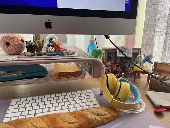 A close-up image of Sendy Santamaria's keyboard and desk with bright yellow headphones, small whimsical objects and toys on a little shelf, and a soft wrist rest that looks like a French baguette.