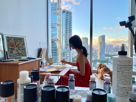 Mae Waite in her high-rise painting studio, with views of other buildings and a sunset visible through the large windows.