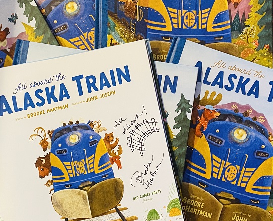 An image of several copies of of All Aboard the Alaska Train. The top copy is open to the title page, which is signed by the author, Brooke Hartman with a picture of a train track.

