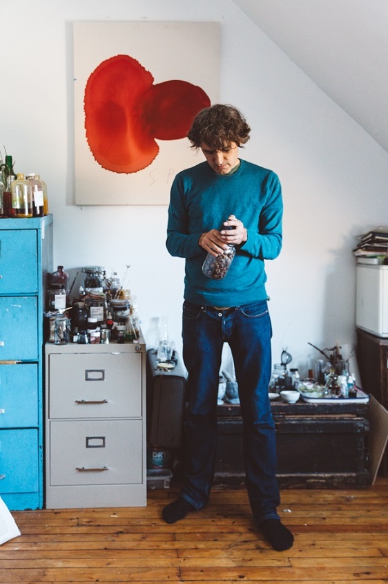 Jason Logan in his studio with a jar of natural materials that appear to be acorns. A painting above his head shows ink swatches made from a beautiful red color.