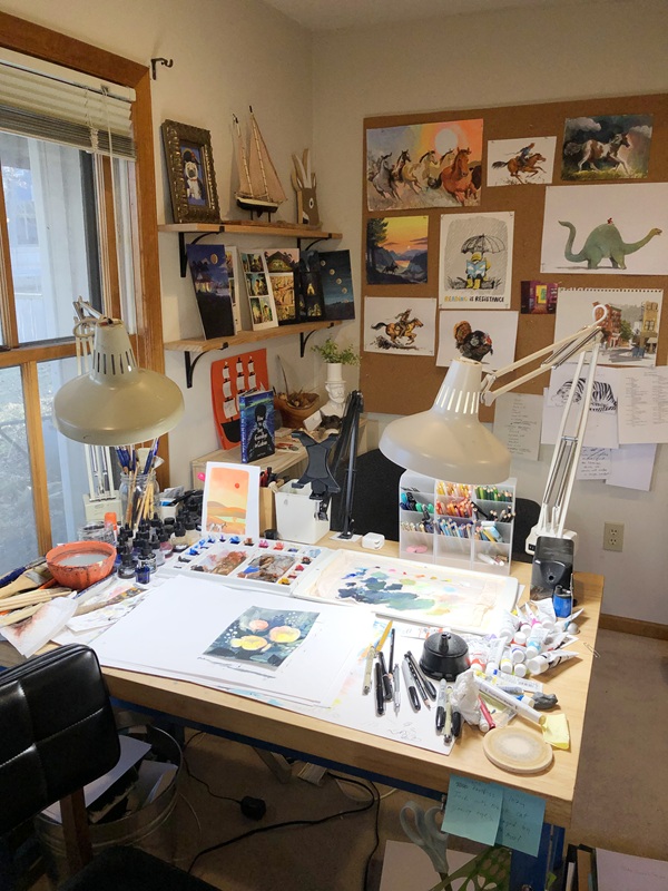 Daniel Miyares' studio, with a drafting table filled with paints and supplies, and a wall where finished artworks are pinned up on display.